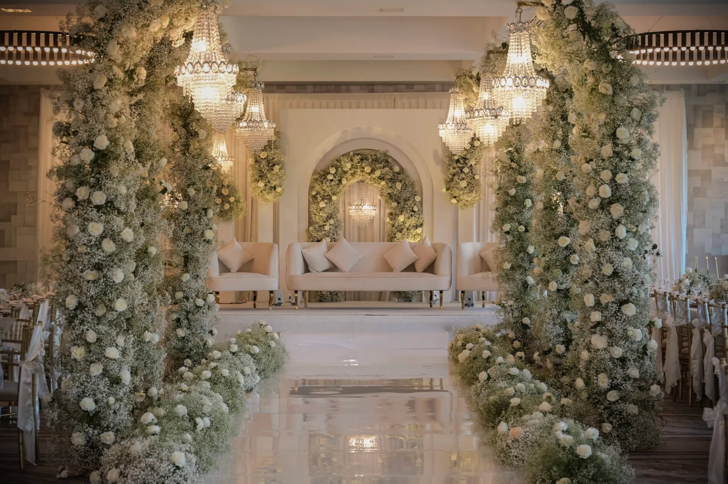A walk-through tunnel of ivory roses and greenery under crystal chandeliers, leading to a white stage