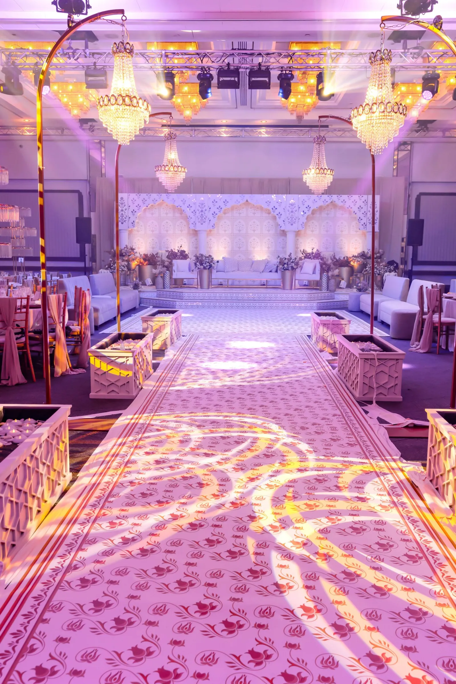 A patterned aisle lit in lilac under chandeliers, leading to a white lattice stage