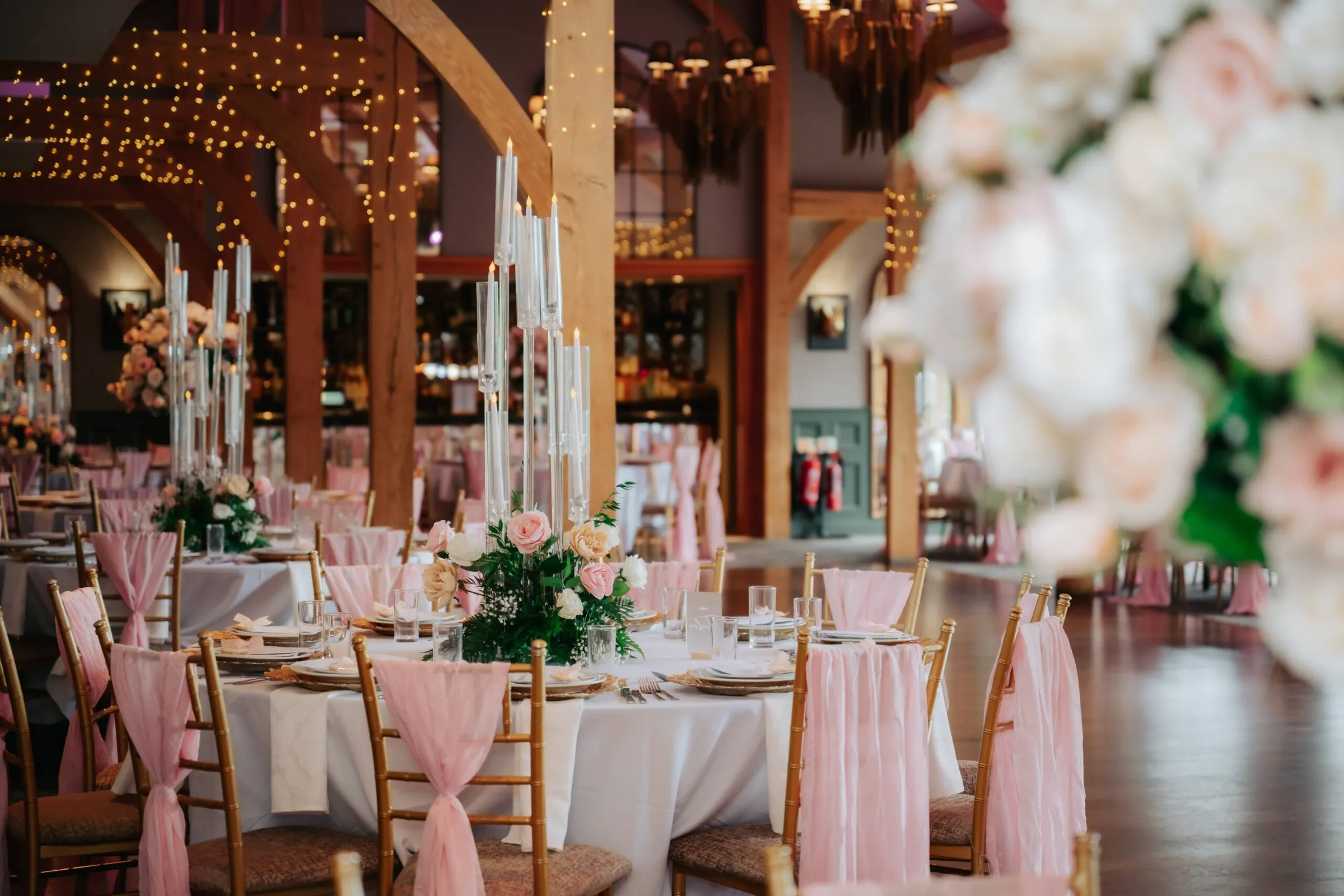 Wedding reception tables dressed in blush and white with tall candles and roses, in a timber-beamed hall