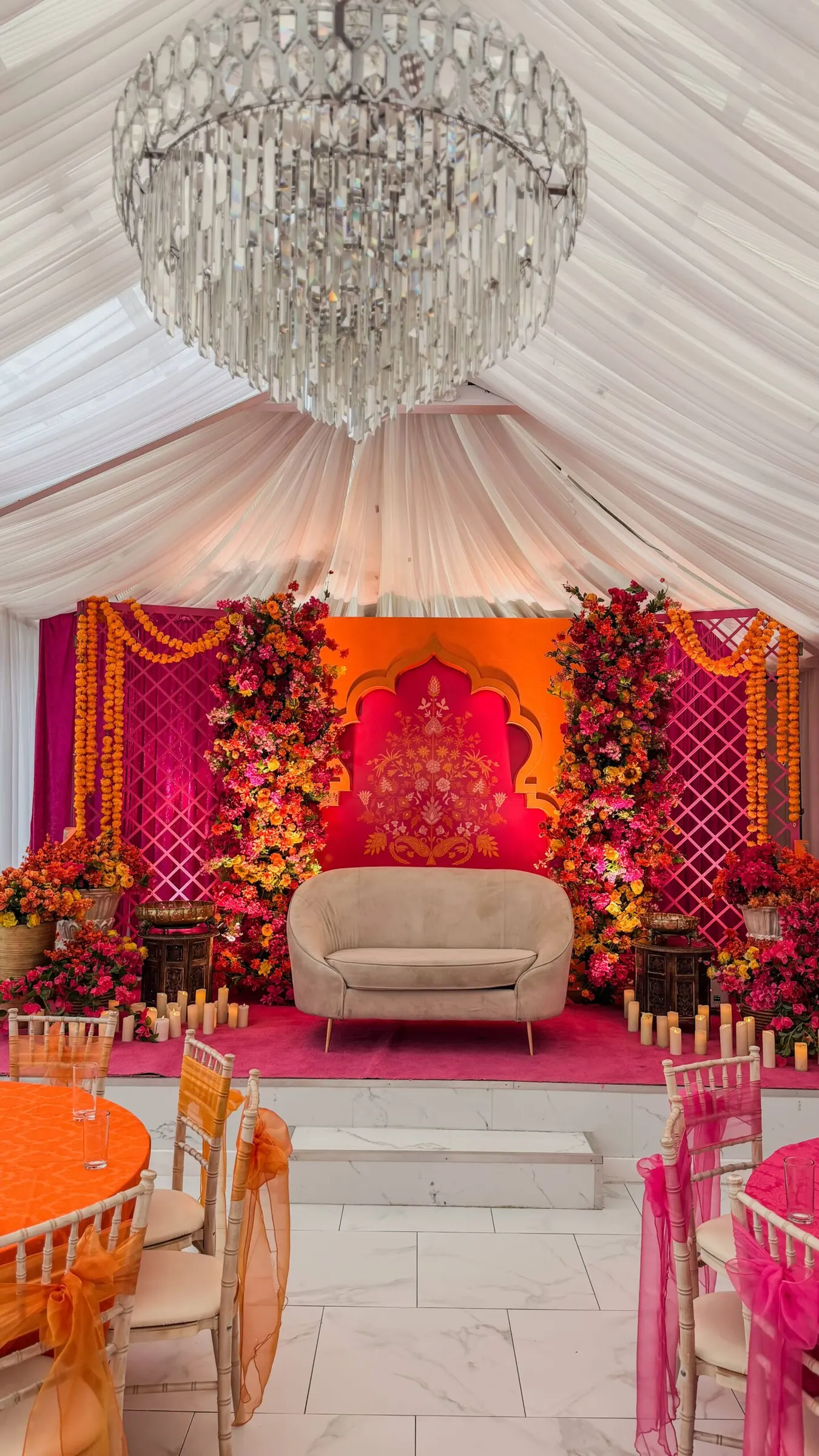 A Mehndi stage in vivid pink, orange and red beneath a crystal chandelier and ivory ceiling drapes