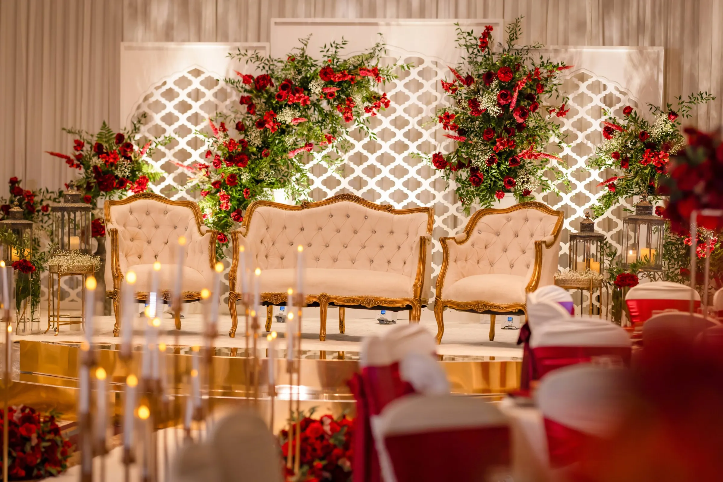 A red and gold wedding stage by Diamond Events: tufted gold settees beneath red rose arrangements and a white lattice