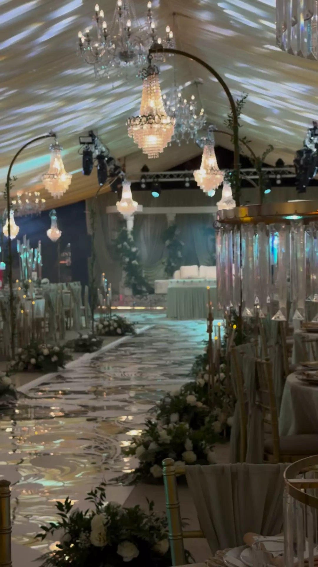 A Diamond Events wedding stage: white columns wrapped in greenery and white flowers, sage drapes and an ivory settee, with a lit aisle leading to it